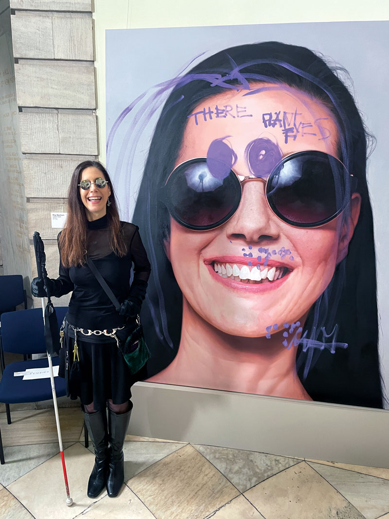 a young woman in sunglasses holding a cane stands in front of a poster with her image